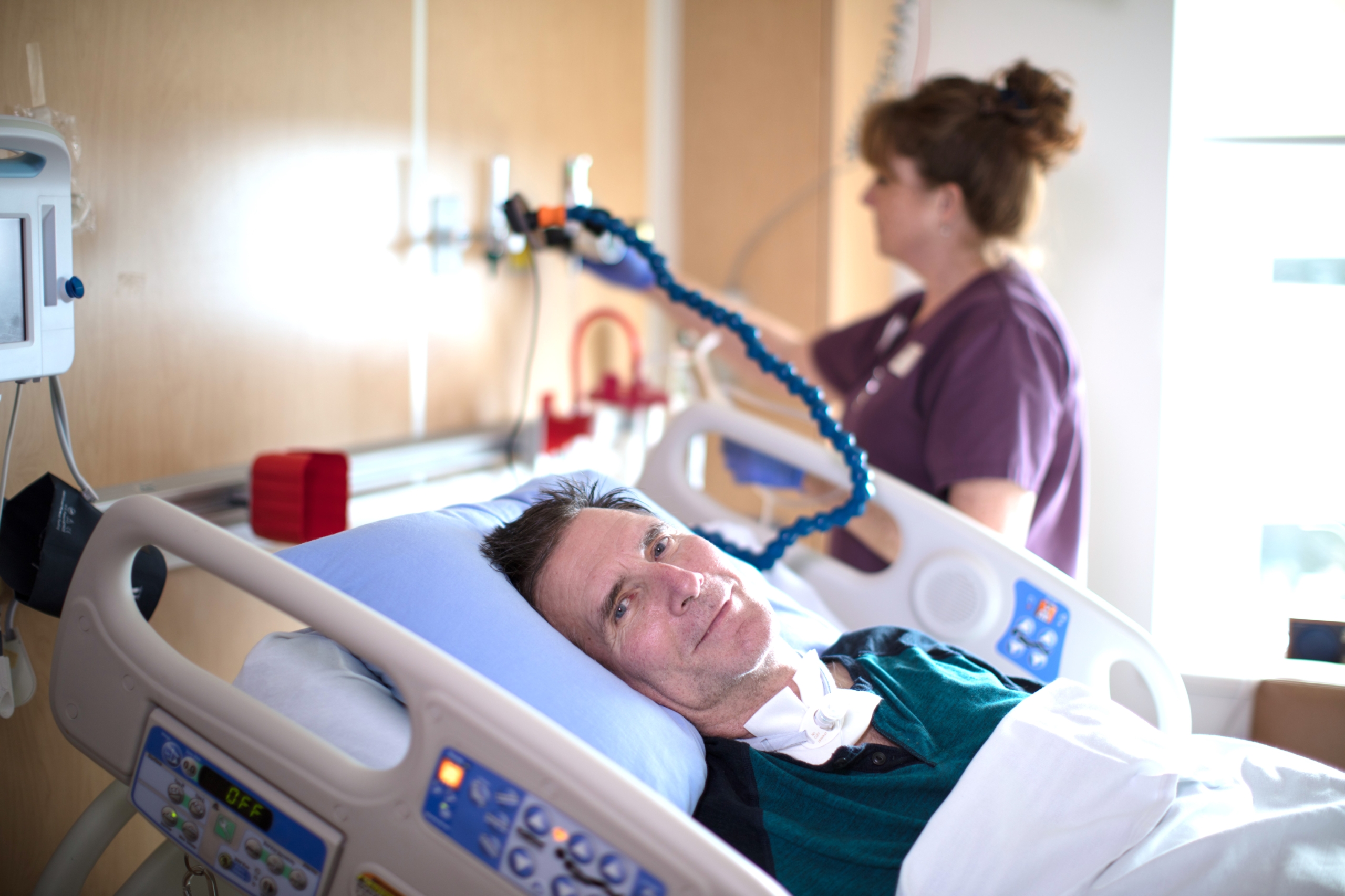 Patient with a tracheotomy looks at the camera while a nurse works in the background
